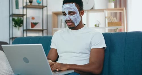 Man Uses Laptop With Facial Mask On At Home