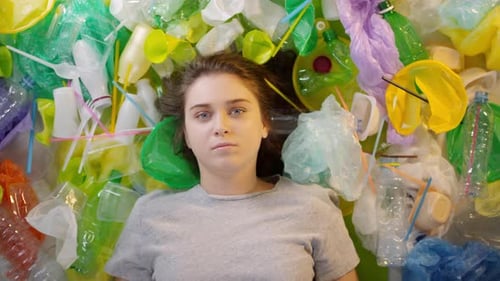 Young Woman Lying Amongst Plastic Waste Holding Sign