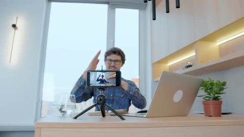Young blogger writes a video for a blog using a smartphone while sitting at a laptop at home.