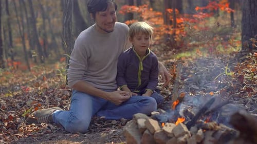 Young Man and His Little Son Sitting By a Bonfire in a Cold Autumn Forest. Autumn Vacation Concept