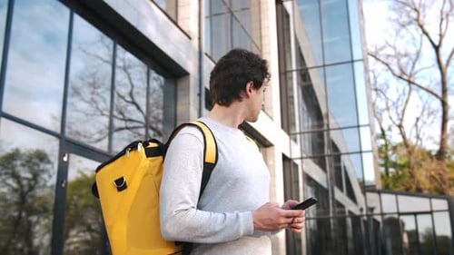 Porträt eines kaukasischen Zustellers, der mit gelbem Rucksack steht, mithilfe der Navigations-App am Telefon