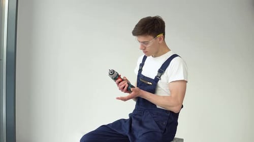 Young Craftsman in Work Uniform and with a Screwdriver in His Hands Sits on a White Wall Background