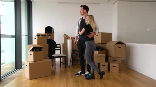 A Smiling Moving Couple Looks Out a Window in a New Apartment - Piles of Cardboard Boxes