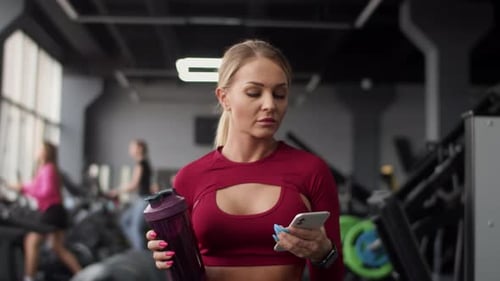Beautiful Athletic Woman with Headphones with Bottle in Hand Walks Through Gym to Treadmill for