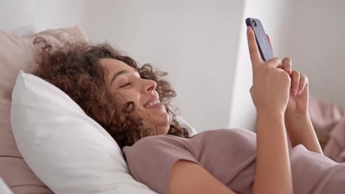 Woman Lies in Bed Laughing at Cell Phone