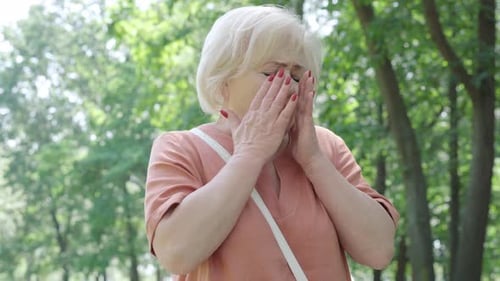 Senior Allergic Woman Sneezing in Summer or Spring Park with Pollen Flying Around. Female Caucasian