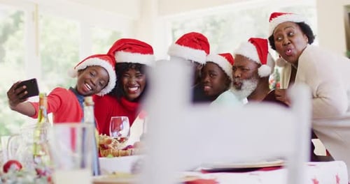 African american family in santa hats taking a selfie on smartphone while sitting on dining table ha