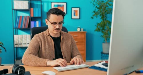 Handsome Brunet with Stubble and Glasses Sits in a Corporate Office at a Desk in