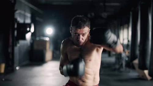A Muscular Sportsman is Boxing in the Dark Gym