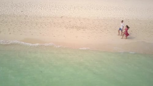Aerial view of young caucasian couple walking hand in hand on white sandy beach in Asia - static cam