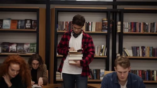 Slow Motion Footage of a Guy in Plaid Shirt and Headphones Walking By Row with Pile of Books and