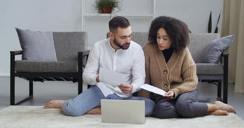 Multi Ethnic Couple Sitting at Home with Laptop Holding Papers Reading Tax Notice Get Loan From Bank