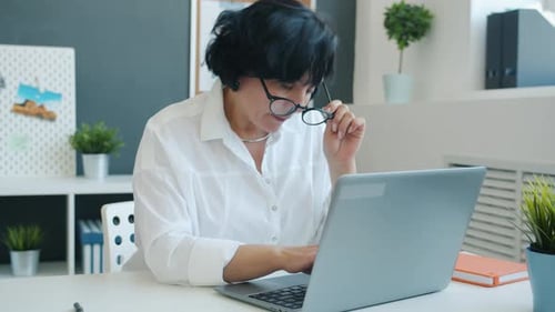 Mature Office Worker Using Laptop Typing Then Taking Off Glasses and Touching Head at Work