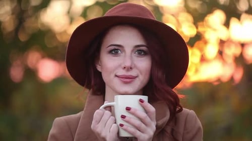 Redhead girl in yellow hat and coat with scarf and cup of coffee on autumn park