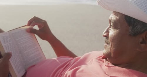 Profile of hispanic senior man relaxing on sunbed on beach at sunset, reading book