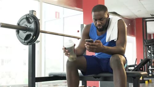 Sportsman Sitting at the Gym, Drinking Protein Cocktail and Scrolling on Phone