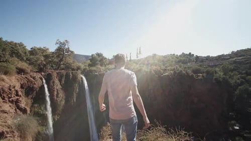 SLOW MOTION: YOUNG CONFIDENT MEN RUNNING TOWARDS EDGE OF CANYON CLIFF WITH WATERFALL SUNNY DAY