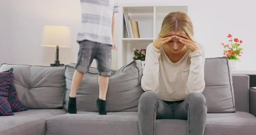 Tired Mother Covering Ears Having Headache While Son Jumping On Couch At Home Parenting Hyperactive