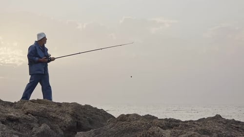 Old fisherman standing on sea side rocks and fishing against the sunset