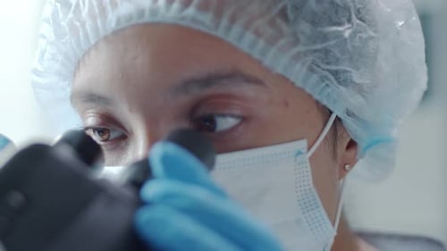 Wissenschaftlerin in medizinischer Maske, Handschuhen und Hut blickt durchs Mikroskop