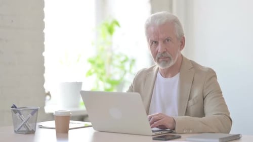 Old Man Looking at Camera While Using Laptop in Office