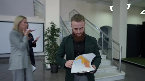 Concentrated Bearded Business Analyst Walking Analyzing Paperwork with Colleagues Passing in Office