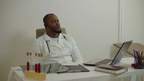Selbstbewusster afrikanischer männlicher Arzt im drahtlosen Headset, der Patienten mithilfe von Video-Chat