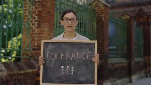 Young Female Teacher Standing Looking Down and at Camera Holding Board with Message Tolerance on
