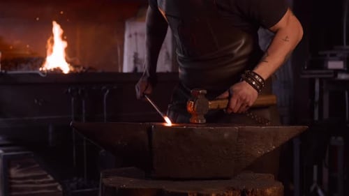 Focused young blacksmith heating and striking metal on anvil in workshop