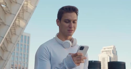 Young Sportsman with Headphones Using Smartphone and Smiling While Standing Outdoors at Stadium