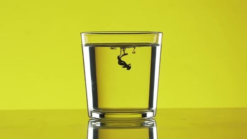 Two Small Drops of Black Ink Dripping Into Clear Water in Glass Standing on Yellow Background in