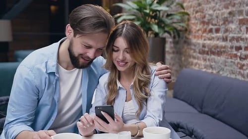 Young Couple Hugging and Watching at Woman's Phone while Sitting at the Table in Hotel Lobby