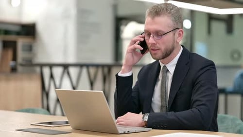 Hardworking Businessman with Laptop Talking on Smartphone in Office