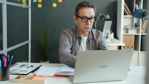 Mature Office Worker Using Laptop Speaking To Himself Then Taking Notes in Notebook at Work