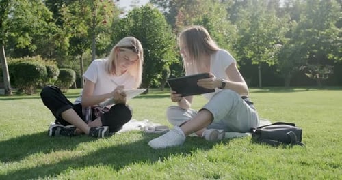 Girls Teenage Students Sitting on Green Lawn in Park with Backpack, Books, Digital Tablet