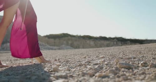 Lady in Blowing Dress and Shoes with Bow in Hand Walks on Beach Stones