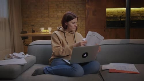 30s Woman Working with Paper Documents and Laptop Sitting on Couch at Home in the Evening