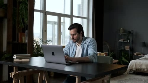 A man Works on a Computer laptop Online, Sitting at a Table at Home in the Evening in a Dark Room