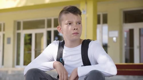 A Caucasian Teenage Boy Looks Around As He Sits in Front of School Closeup