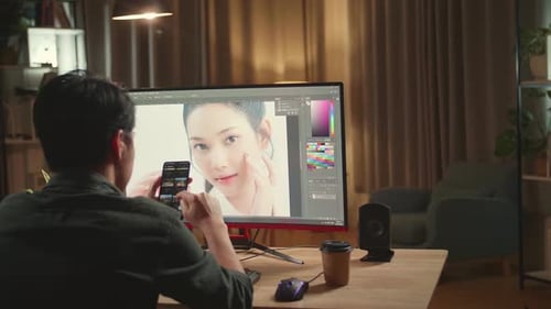 Asian Male Digital Editor Using Mobile Phone While Works In Photo Editing Software On Computer