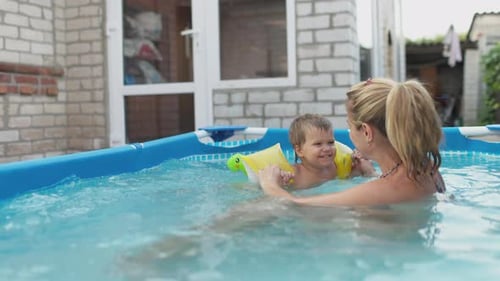 Mama spielt mit einem nackten Baby in Überärmeln im Pool vor dem Hintergrund eines Sommersonnenuntergangs