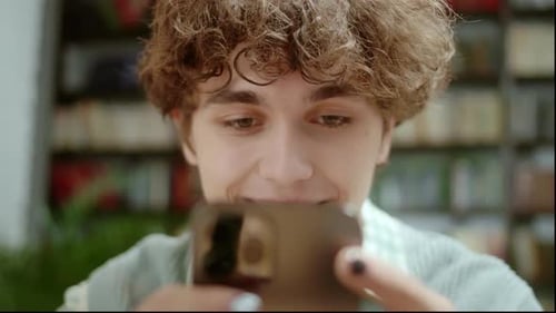 Handsome Curly Student Man Holding Modern Smartphone Texting Message in Home Office or Library