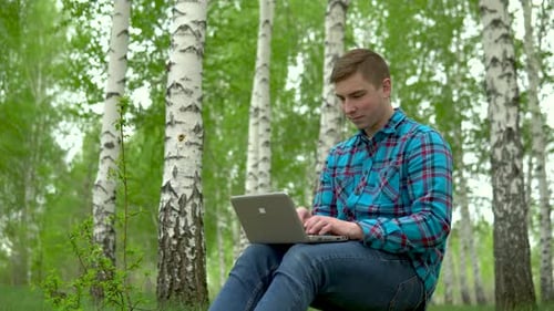 Young Man in Nature with a Laptop in His Hands. A Man Sits on a Stump in a Birch Forest and Leads