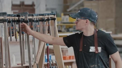 Man Walks in a Carpentry Workshop and Takes an Instrument Looking in the Camera