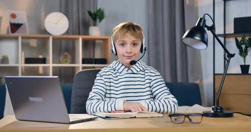 Un colegial inteligente con auriculares que se sienta en su lugar de trabajo durante una lección en línea y mira a la cámara