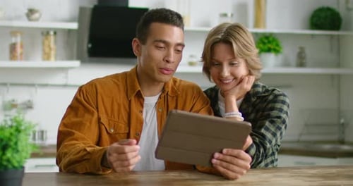 Happy Mixed Race Couple Is Sitting in Their Kitchen, Browsing in Tablet Computer