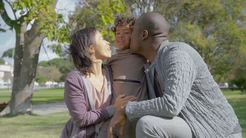 Cheerful Multicultural Parents Hugging and Kissing Son on Green Lawn with Sunlight