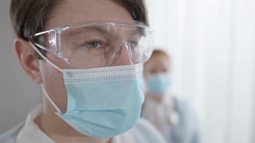 Nahaufnahme Gesicht des kaukasischen Arztes in Covid-19 Gesichtsmaske und Schutzbrille mit verschwommener Krankenschwester