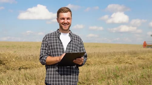 Happy Young Farmer with Notebook Standing on Wheat Field While Combine Harvester Working in