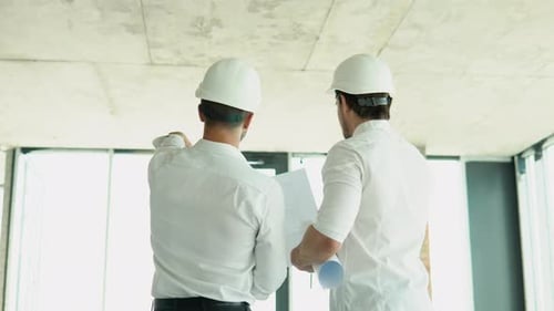 Dois homens de capacete olham para o plano do edifício